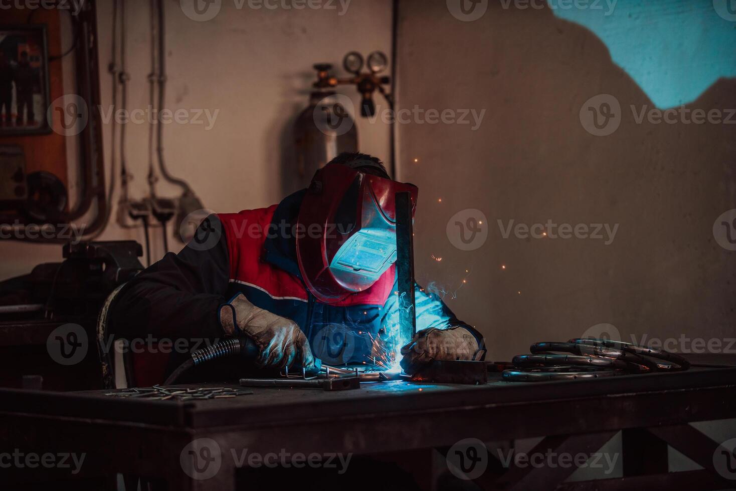 Profesional welder in protective uniform and mask welding metal pipe on the industrial table with other workers behind in the industrial workshop. photo