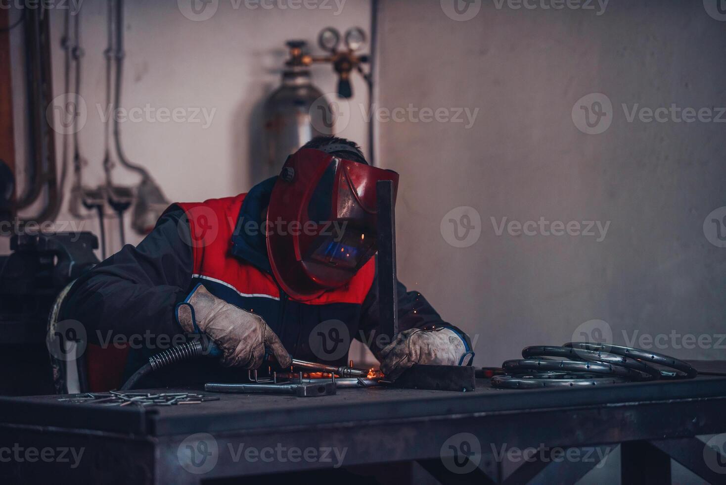 Profesional welder in protective uniform and mask welding metal pipe on the industrial table with other workers behind in the industrial workshop. photo