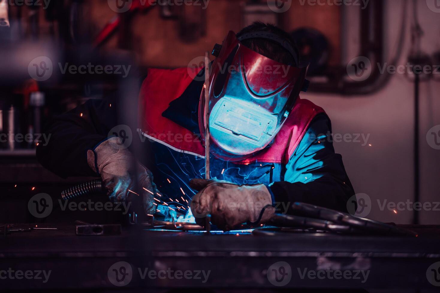 Profesional welder in protective uniform and mask welding metal pipe on the industrial table with other workers behind in the industrial workshop. photo
