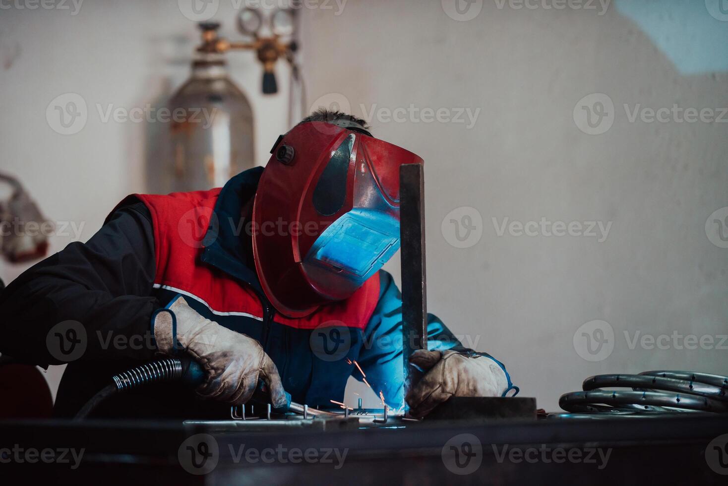 Profesional welder in protective uniform and mask welding metal pipe on the industrial table with other workers behind in the industrial workshop. photo