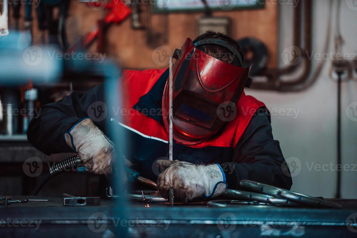 Profesional welder in protective uniform and mask welding metal pipe on the industrial table with other workers behind in the industrial workshop. photo