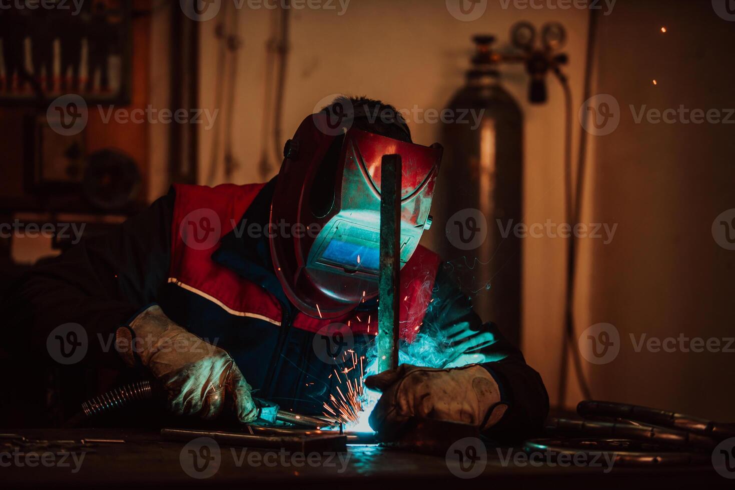Profesional welder in protective uniform and mask welding metal pipe on the industrial table with other workers behind in the industrial workshop. photo