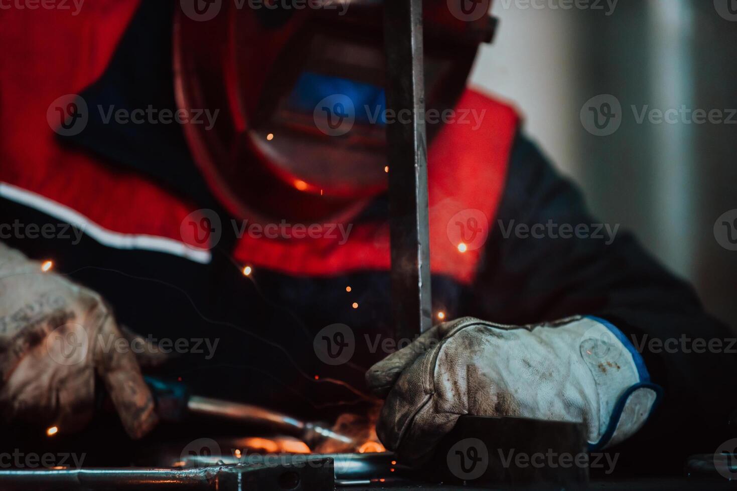 Close up welder in protective uniform and mask welding metal pipe on the industrial table with other workers behind in the industrial workshop. photo