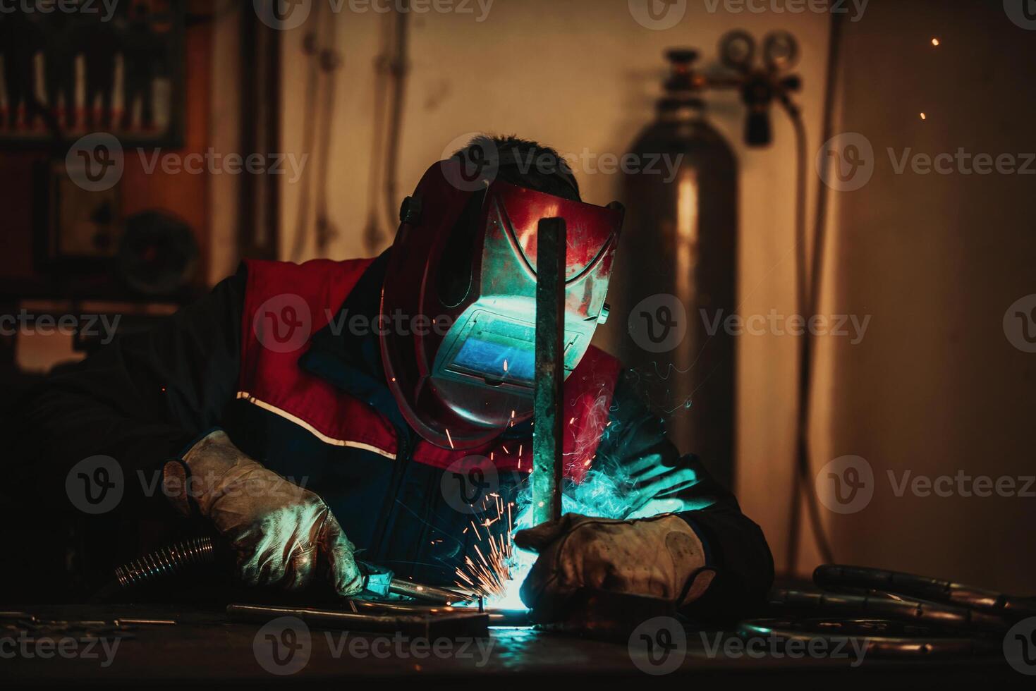 Profesional welder in protective uniform and mask welding metal pipe on the industrial table with other workers behind in the industrial workshop. photo