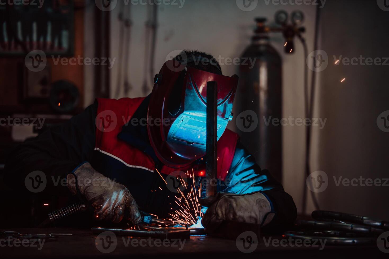 Profesional welder in protective uniform and mask welding metal pipe on the industrial table with other workers behind in the industrial workshop. photo