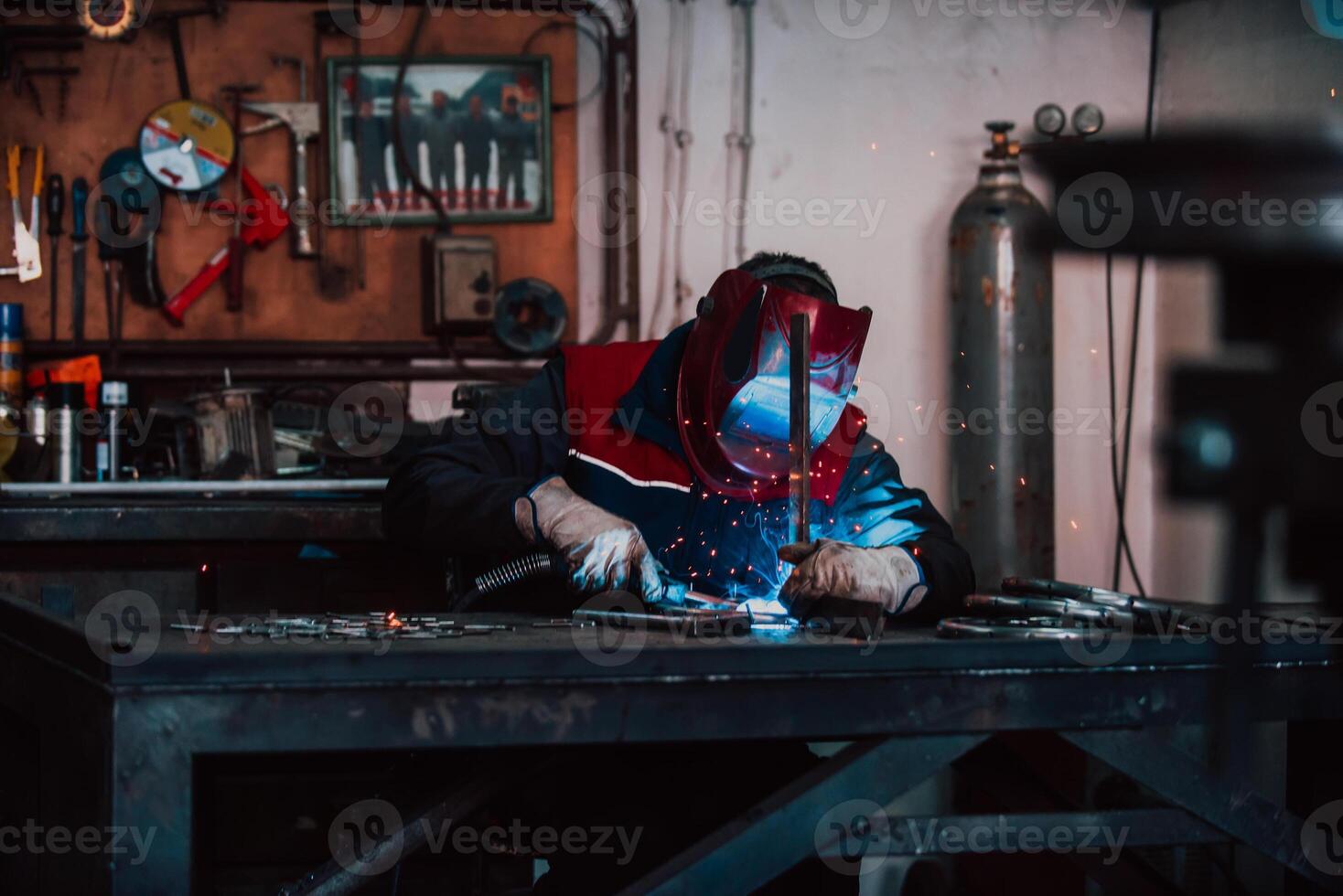 Close up welder in protective uniform and mask welding metal pipe on the industrial table with other workers behind in the industrial workshop. photo
