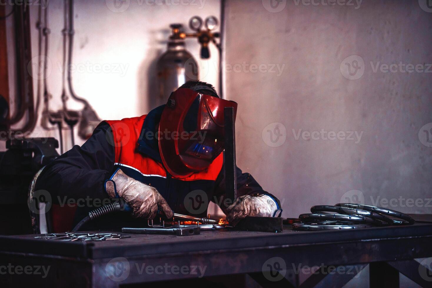 Profesional welder in protective uniform and mask welding metal pipe on the industrial table with other workers behind in the industrial workshop. photo