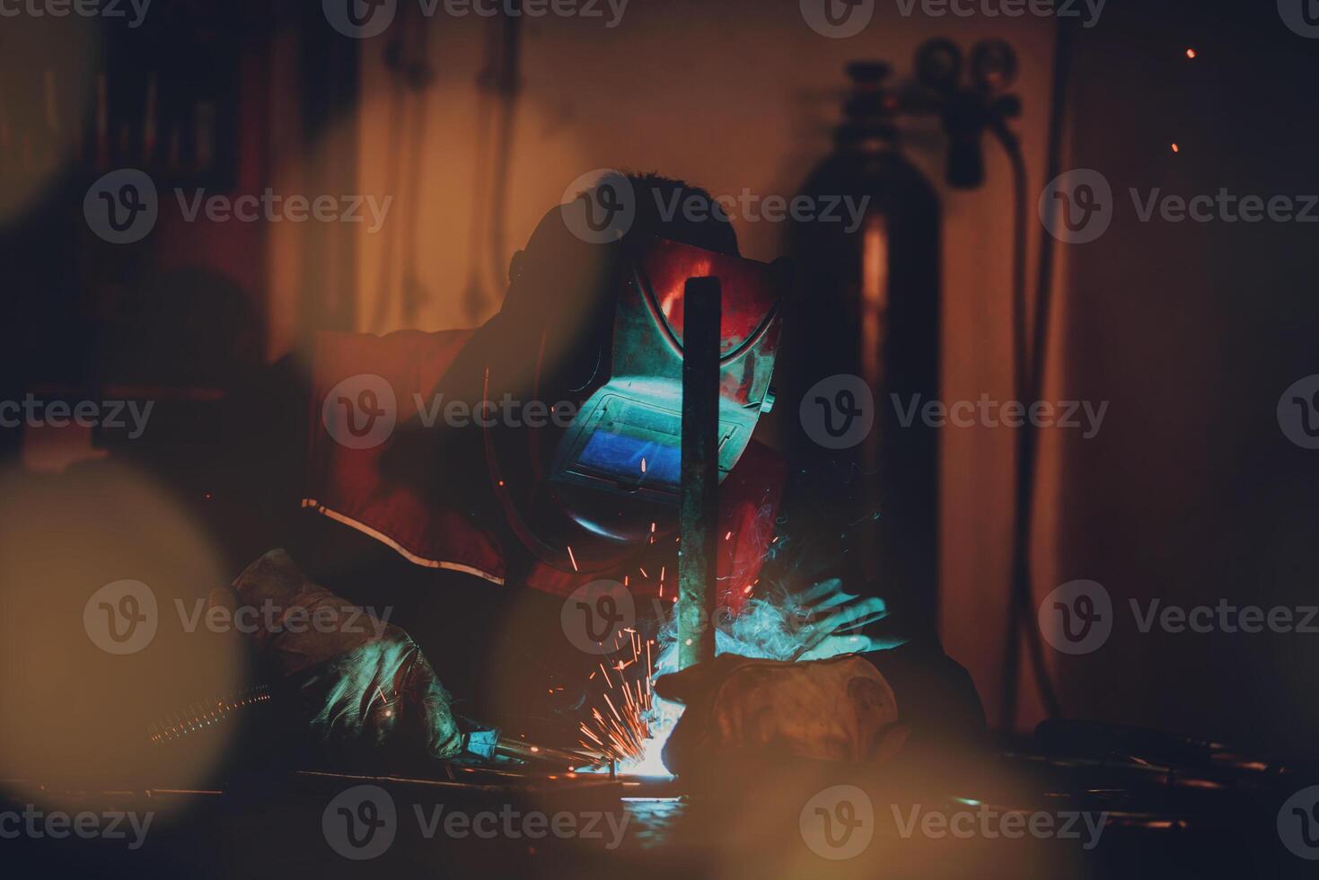 Profesional welder in protective uniform and mask welding metal pipe on the industrial table with other workers behind in the industrial workshop. photo