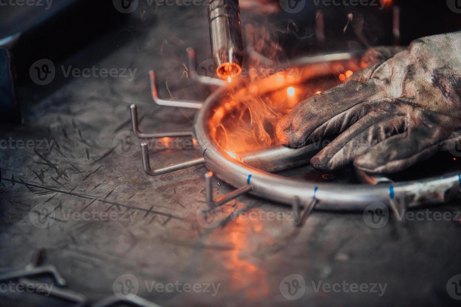 Close up welder in protective uniform and mask welding metal pipe on the industrial table with other workers behind in the industrial workshop. photo