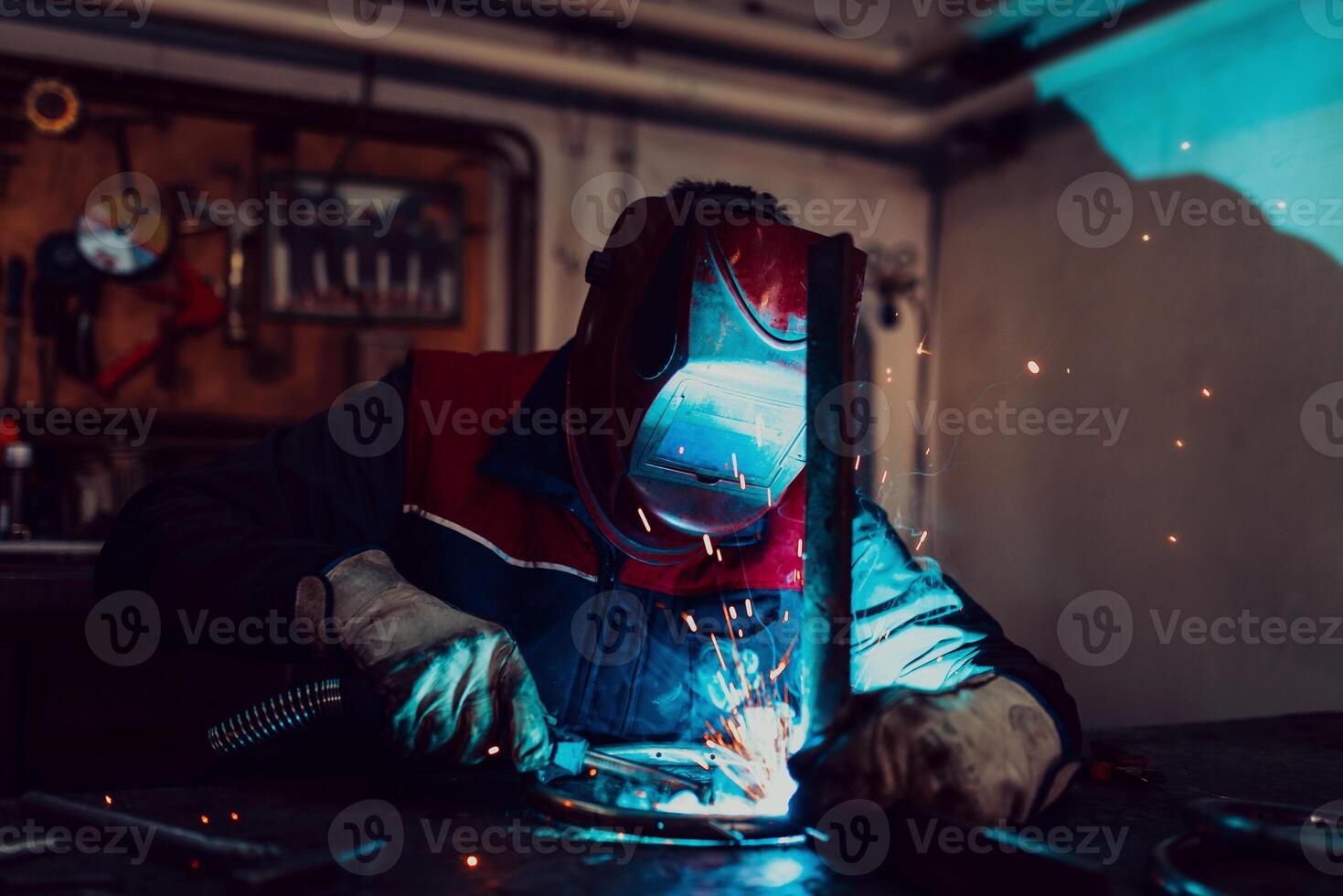 Profesional welder in protective uniform and mask welding metal pipe on the industrial table with other workers behind in the industrial workshop. photo