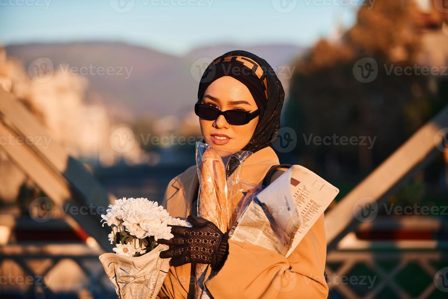 A hijab woman in stylish sunglasses and an elegant French outfit, walking through the city at sunset, carrying a bouquet, bread, and newspaper, radiating a sense of cultural charm and serenity photo