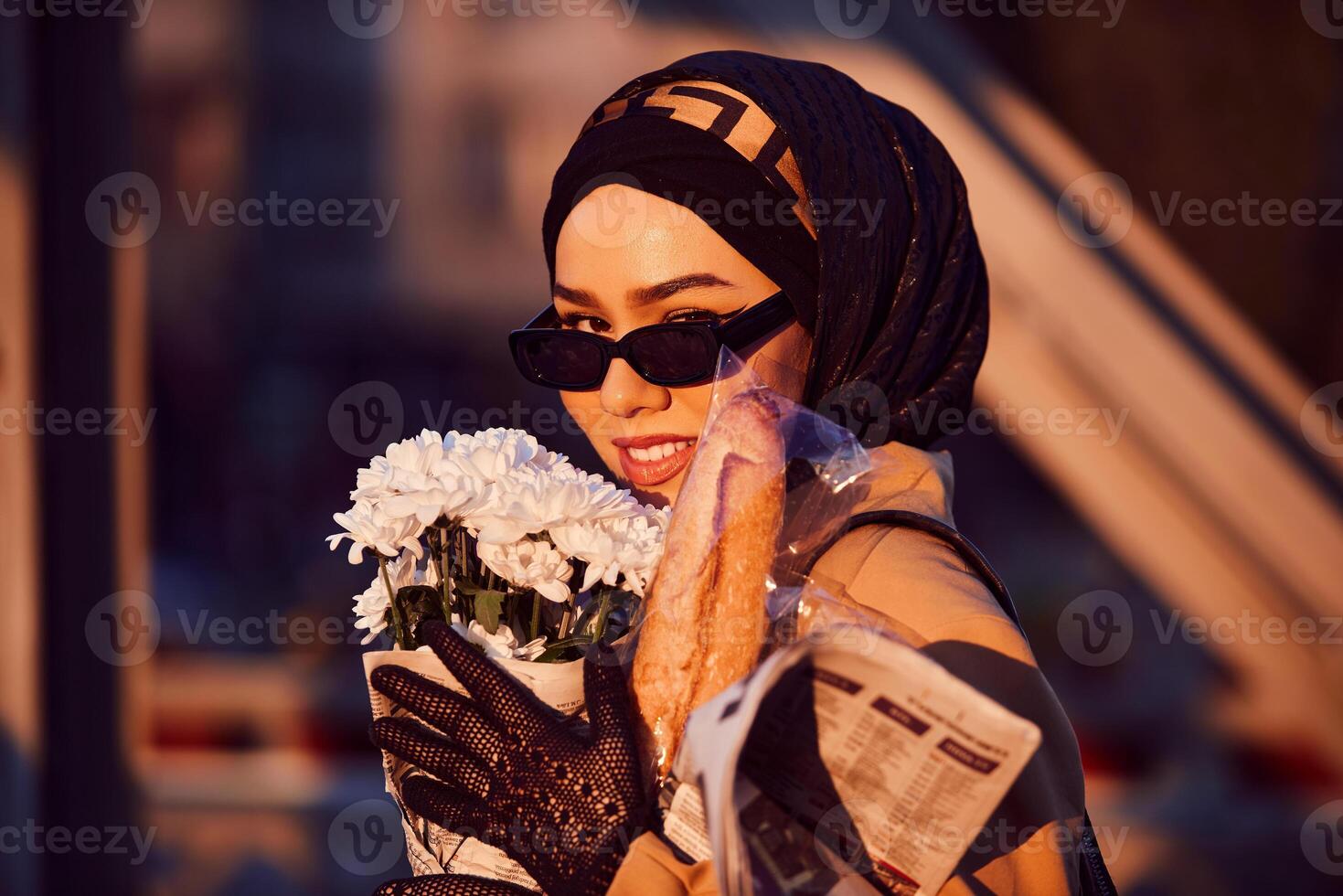 A hijab woman in stylish sunglasses and an elegant French outfit, walking through the city at sunset, carrying a bouquet, bread, and newspaper, radiating a sense of cultural charm and serenity photo