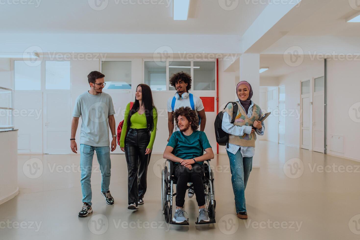 In a modern university, a diverse group of students, including an African American student and a hijab-wearing woman, walk together in the hallway, accompanied by their wheelchair-bound colleague. photo