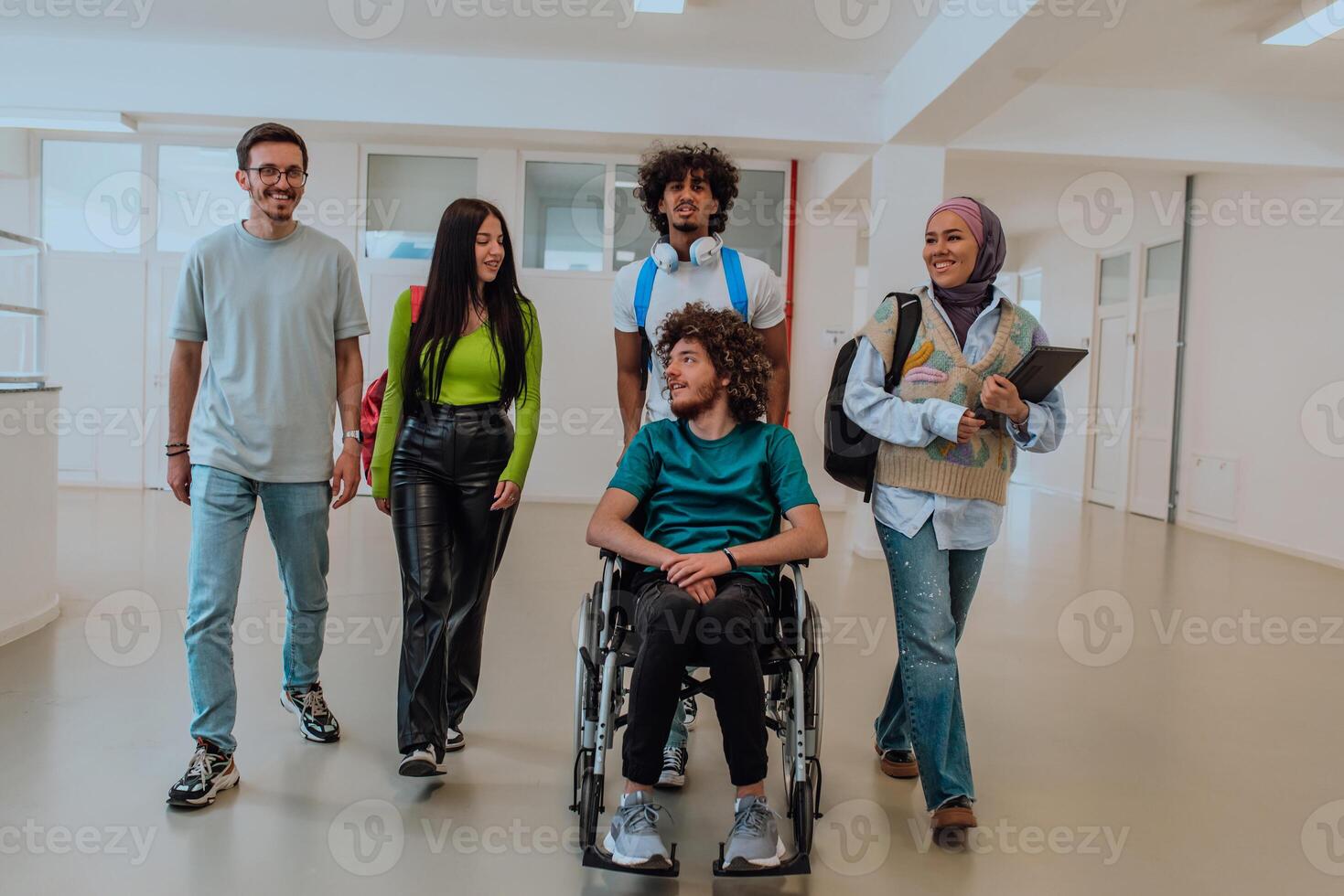 In a modern university, a diverse group of students, including an African American student and a hijab-wearing woman, walk together in the hallway, accompanied by their wheelchair-bound colleague photo