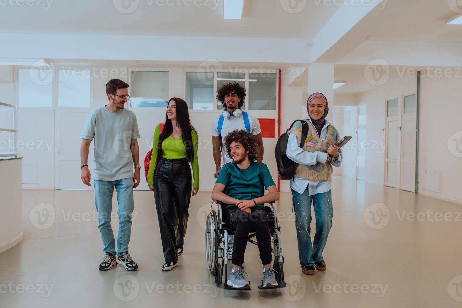 In a modern university, a diverse group of students, including an African American student and a hijab-wearing woman, walk together in the hallway, accompanied by their wheelchair-bound colleague photo