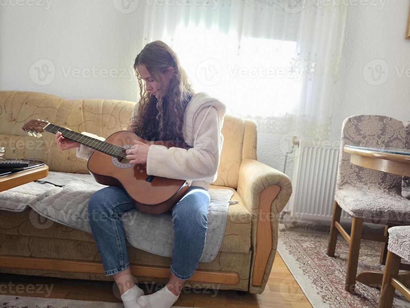 The young, curly-haired girl plays her acoustic guitar with passion and talent in the comfort of her home, creating beautiful melodies in this realistic photograph photo