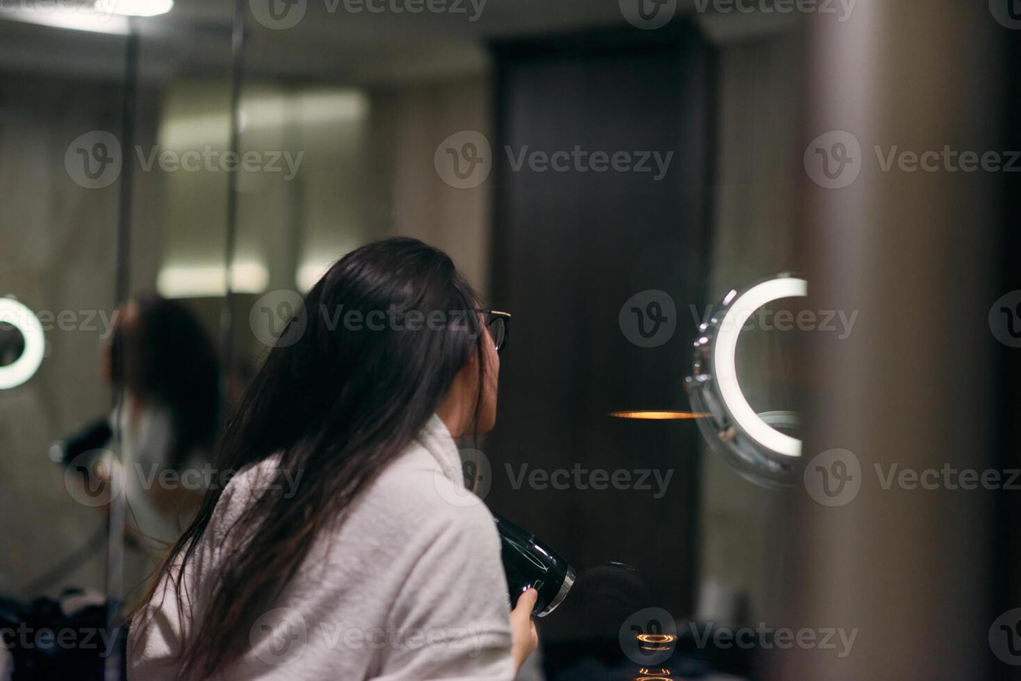 A joyful woman, with headphones in her ears, playfully dries her hair with a hairdryer in the bathroom after a refreshing bath, embodying a carefree and energetic moment of self-care and relaxation photo