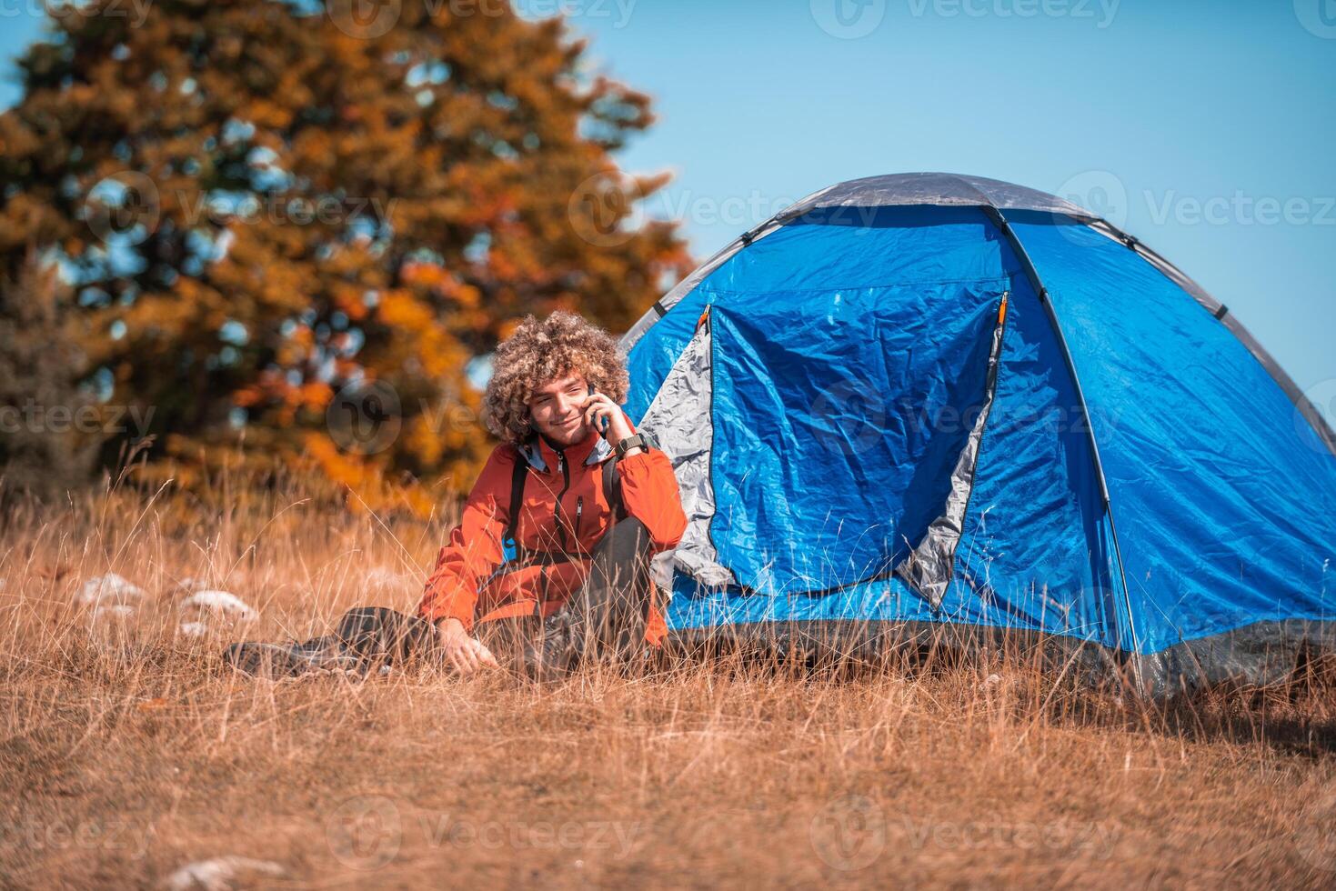 A man with a tent enjoys a sunny day whileusing a smartphone and camping in the mountains photo
