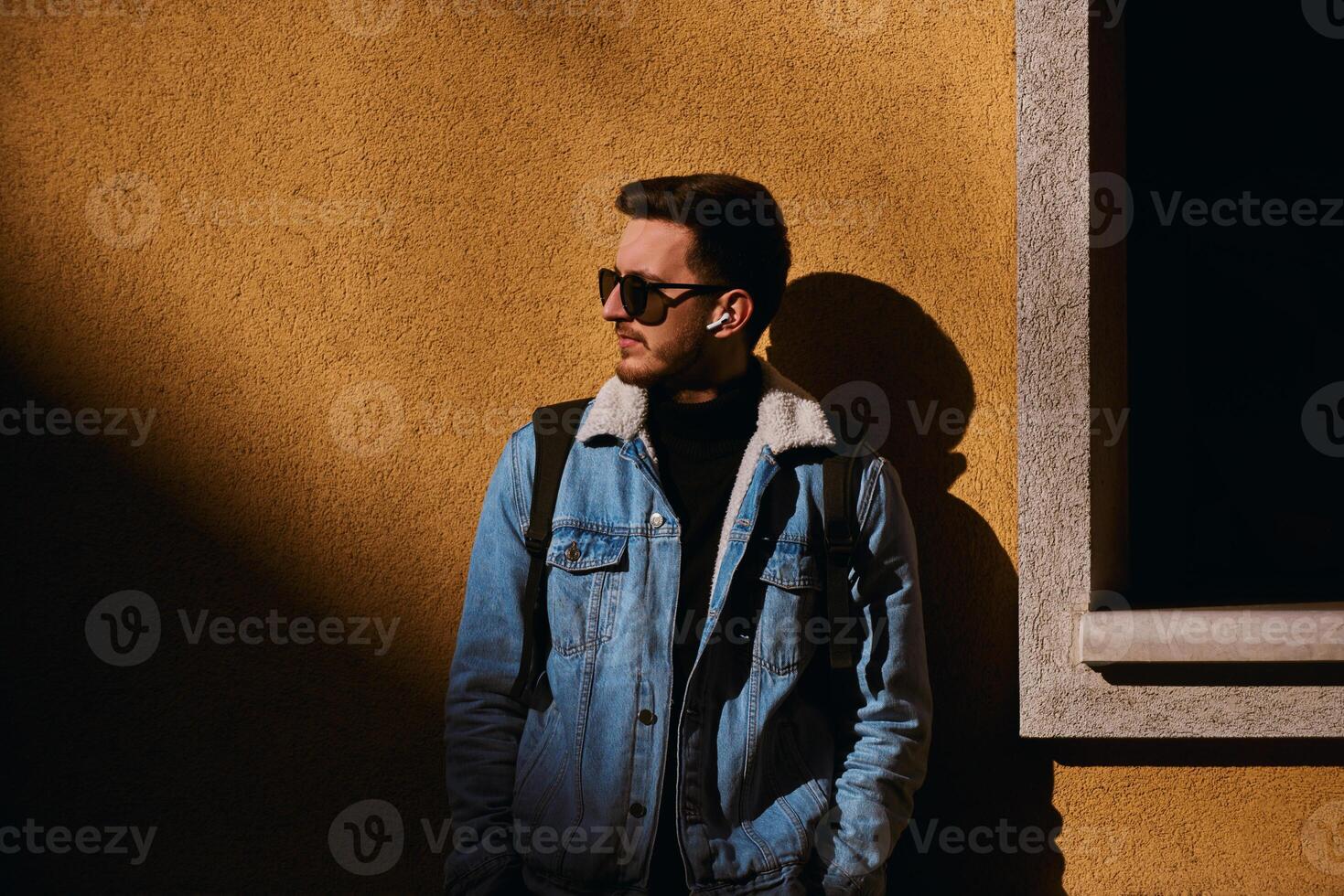 A man stands confidently in front of an orange wall, wearing sunglasses, a backpack, and wireless headphones, exuding a modern urban style and a sense of independence and self-assurance. photo