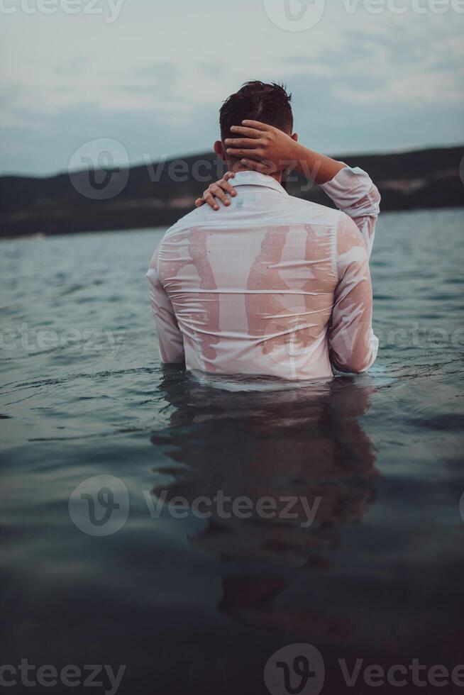 A symbolic and emotionally connected couple tenderly embraces against the backdrop of a serene sea, bathed in the warm hues of a breathtaking sunset photo