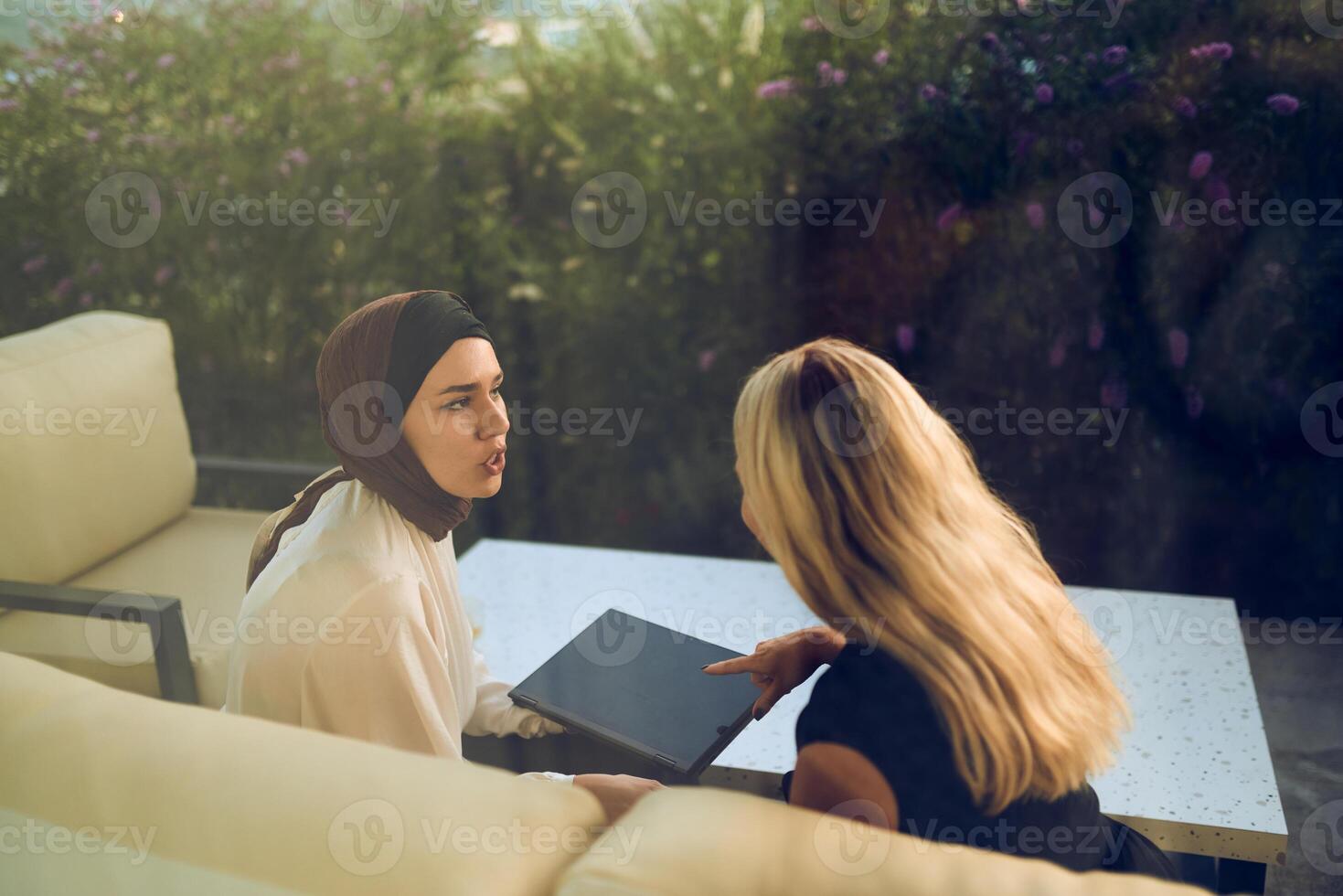 A hijab-wearing woman, a successful real estate business owner, gracefully sells a house to a blonde customer, both sitting in front of the property. photo