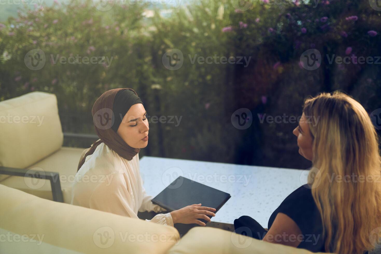 A hijab-wearing woman, a successful real estate business owner, gracefully sells a house to a blonde customer, both sitting in front of the property. photo