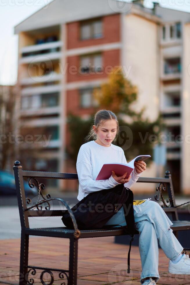 A beautiful blonde teenager sits on a bench in the park, taking a break from school to immerse herself in a book, accompanied by her school backpack photo