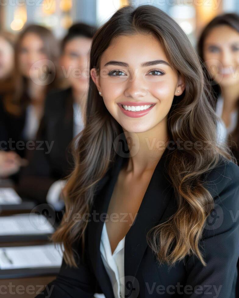 A young woman in a black blazer smiles confidently as she sits in a meeting with other professionals photo