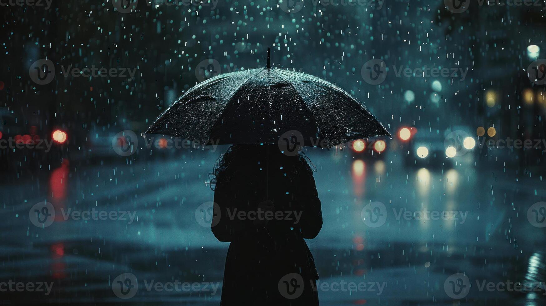 A lone person stands in the rain, holding a black umbrella, with blurred city lights in the background photo