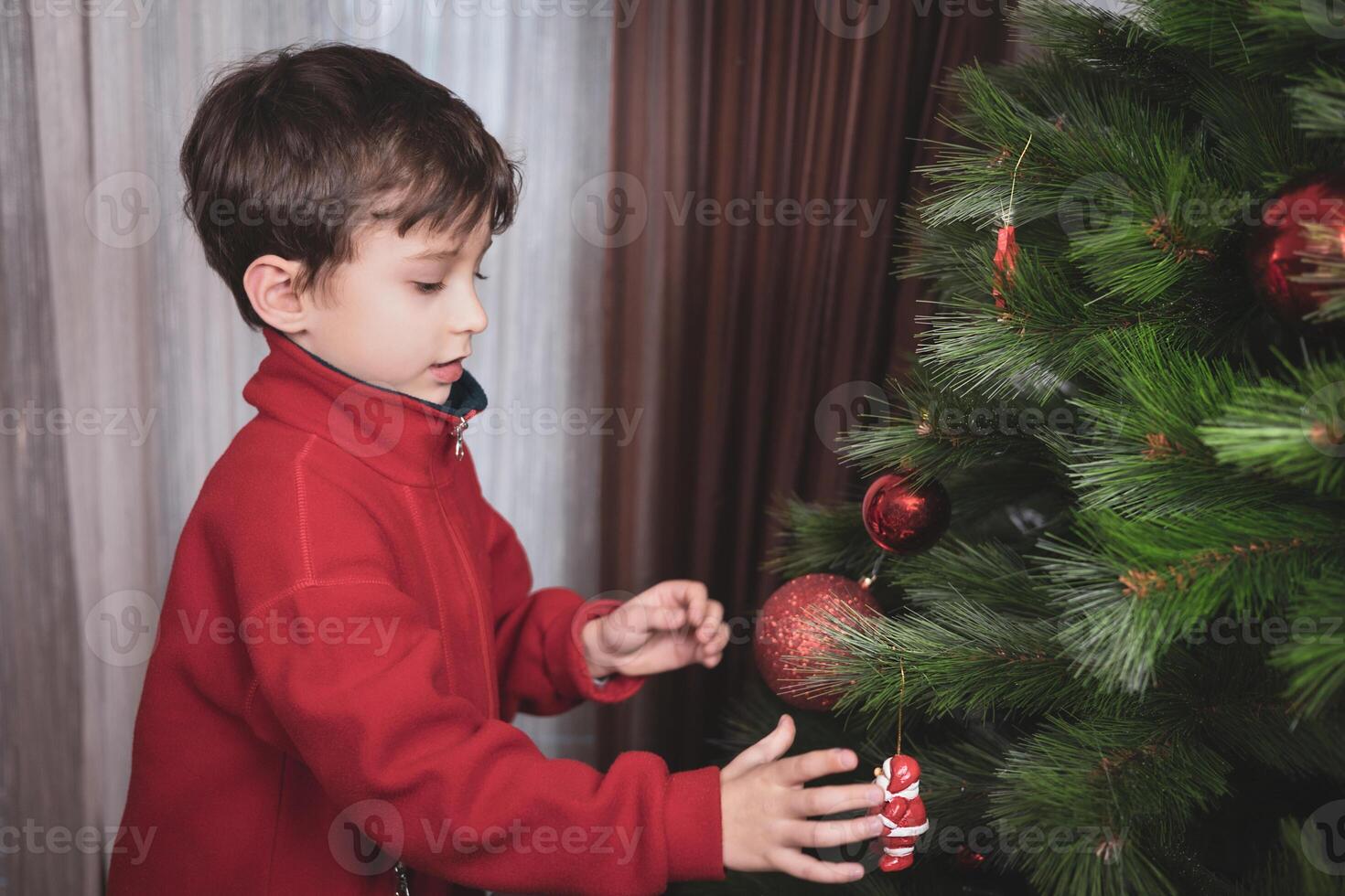 Close-up of a happy little boy decorating the Christmas tree at home with a big glass red ball. photo