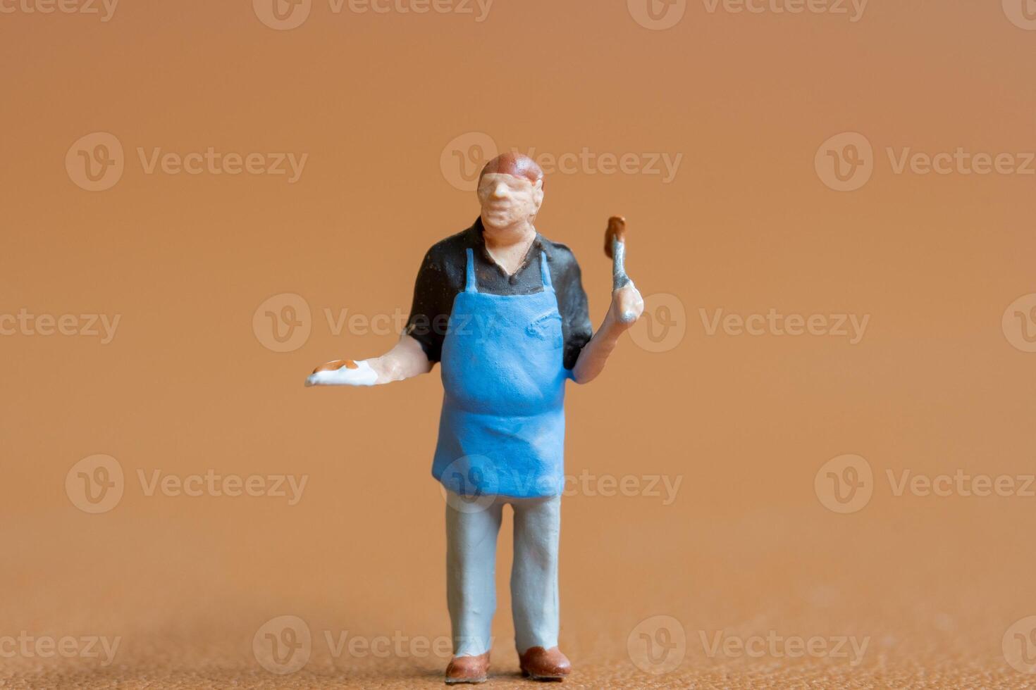 Miniature people , A chef is wearing a blue apron over a black shirt and light-colored pants. The background is a solid, photo