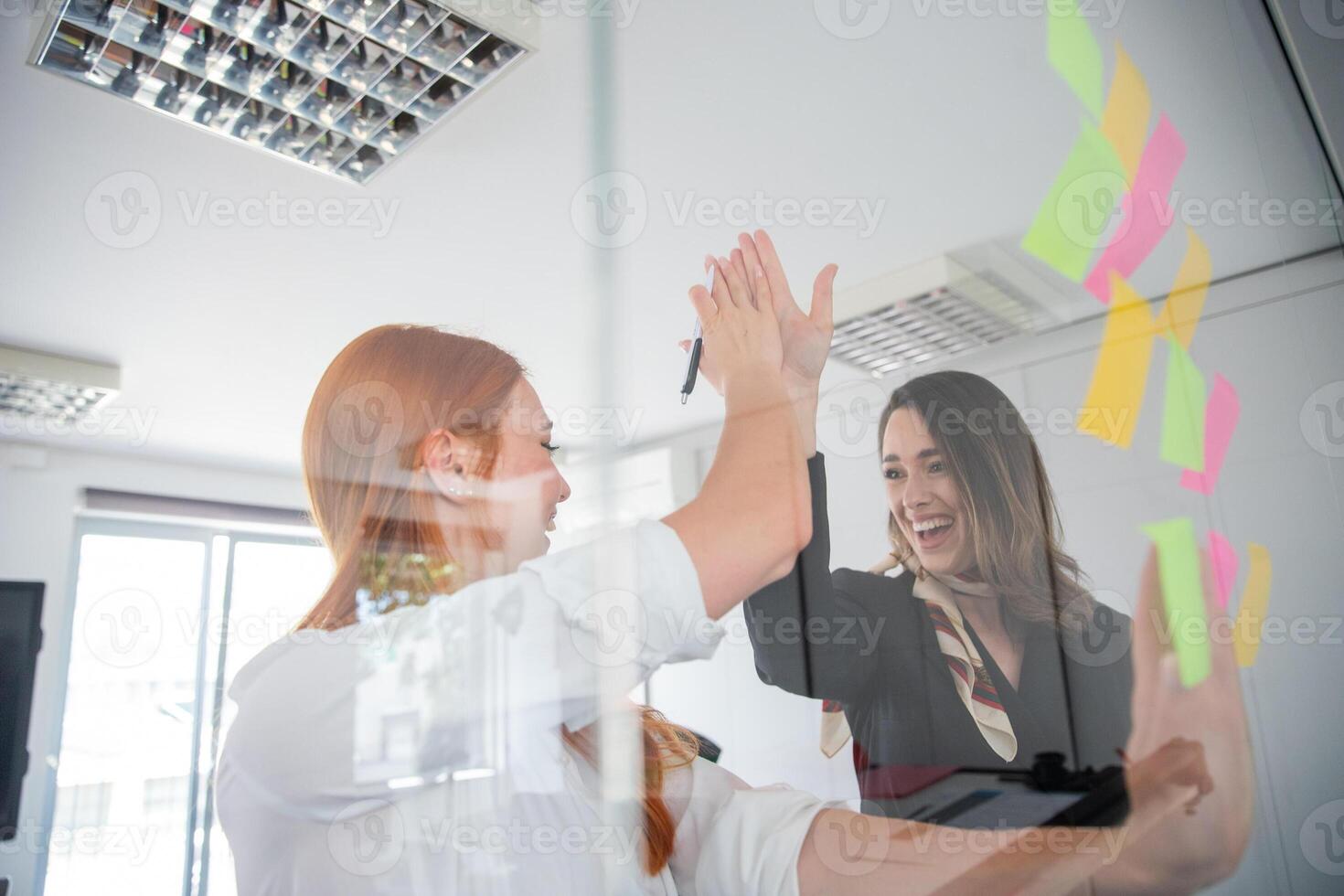 Two colleagues give each other high-fives while writing on stick notes hanging on the wall photo