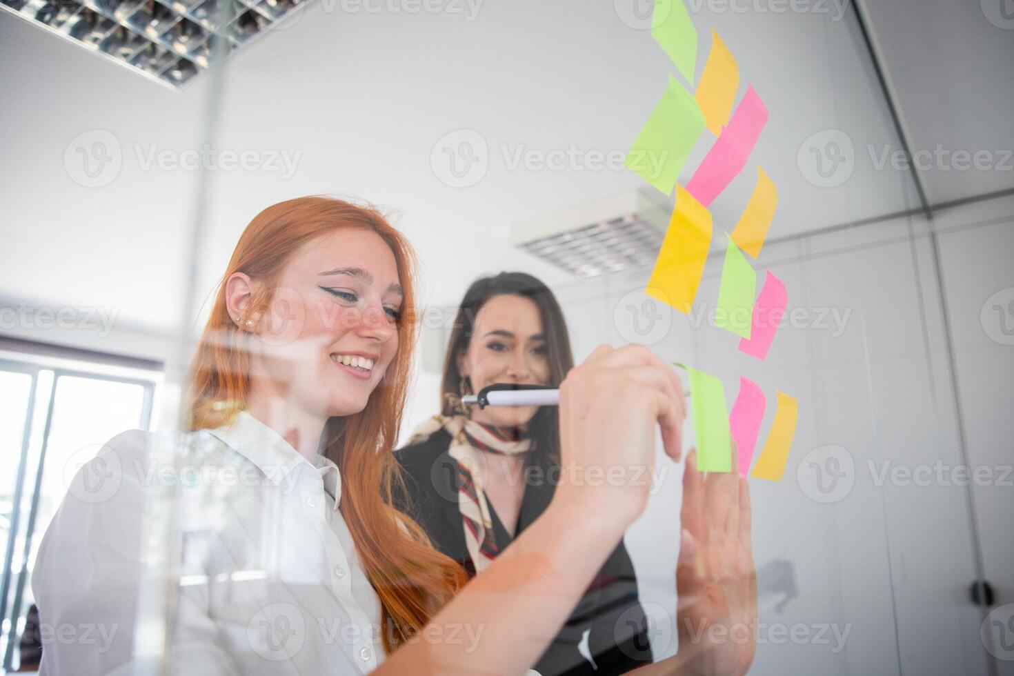 Two businesswomen write on the colored stick notes hanging on the wall during a meeting photo