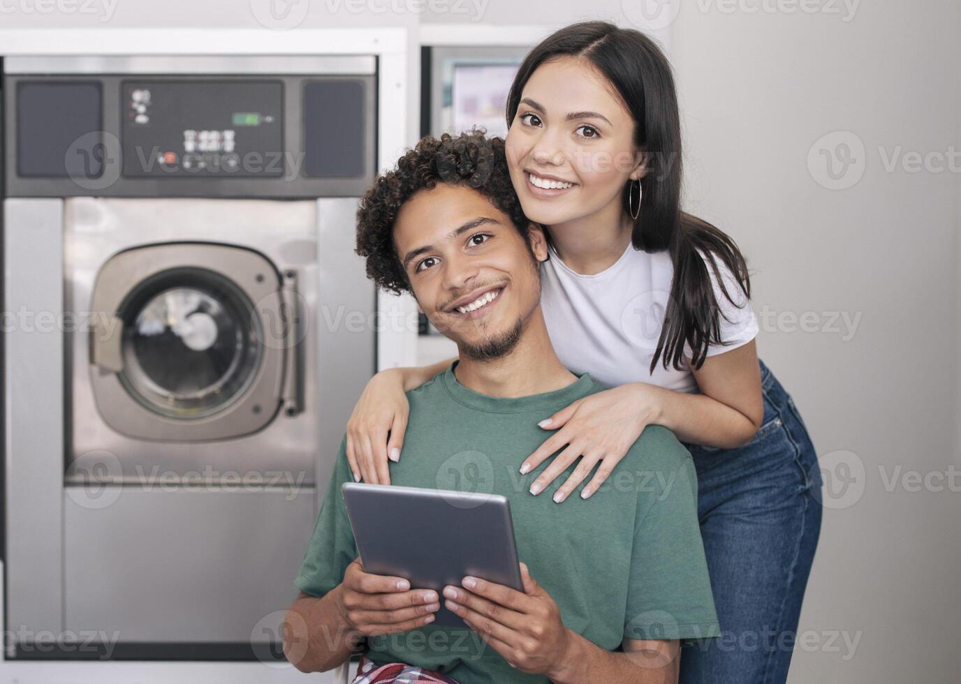 Laundry Service. Happy Young Diverse Couple Posing With Digital Tablet Hugging Near New Washing Machine Indoors, Booking Washer And Smiling To Camera Advertising Laundromat Service photo