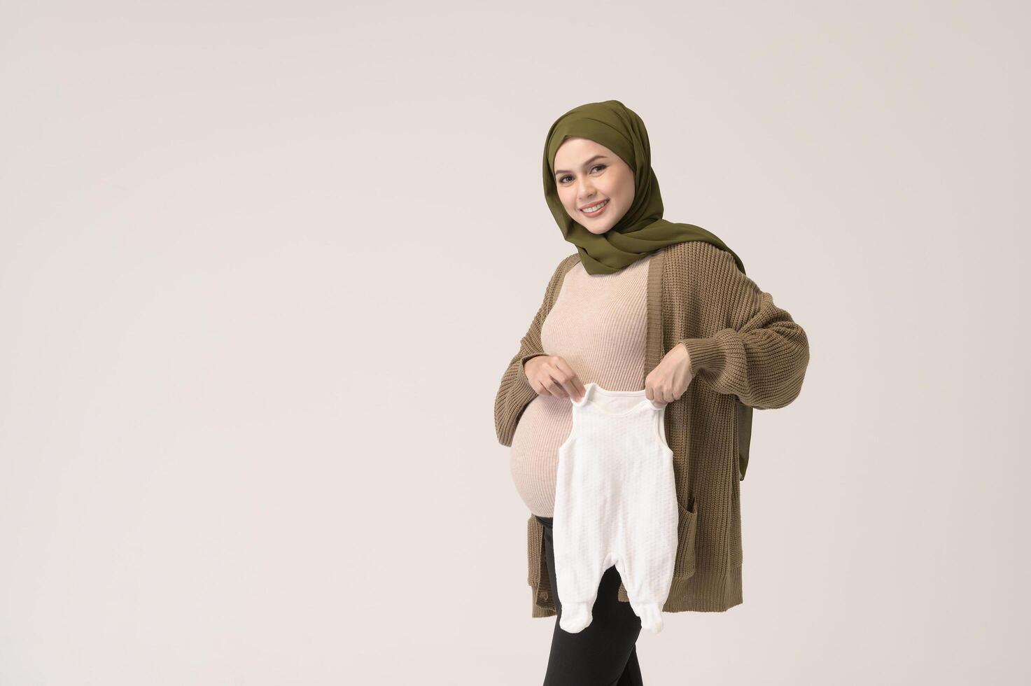 Portrait of Pregnant muslim woman in hijab holding baby cloth over white background studio photo
