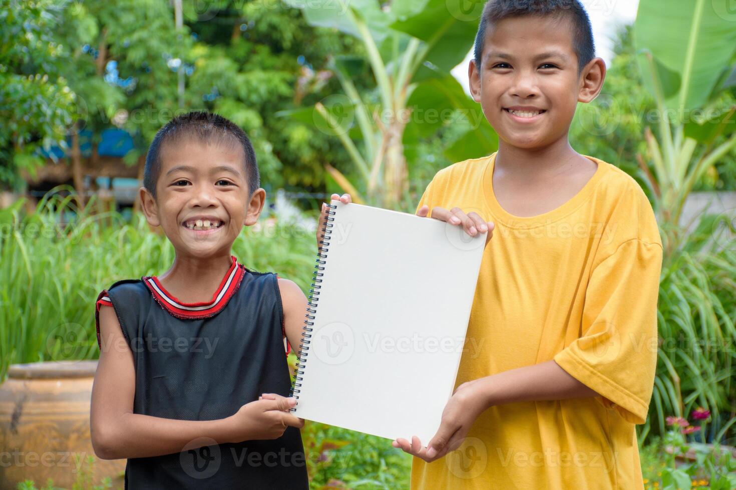 Two boys are smiling charmingly and holding a blank piece of paper or a sign on which to add words or letters in natural forest background at garden for concept of billboard. photo