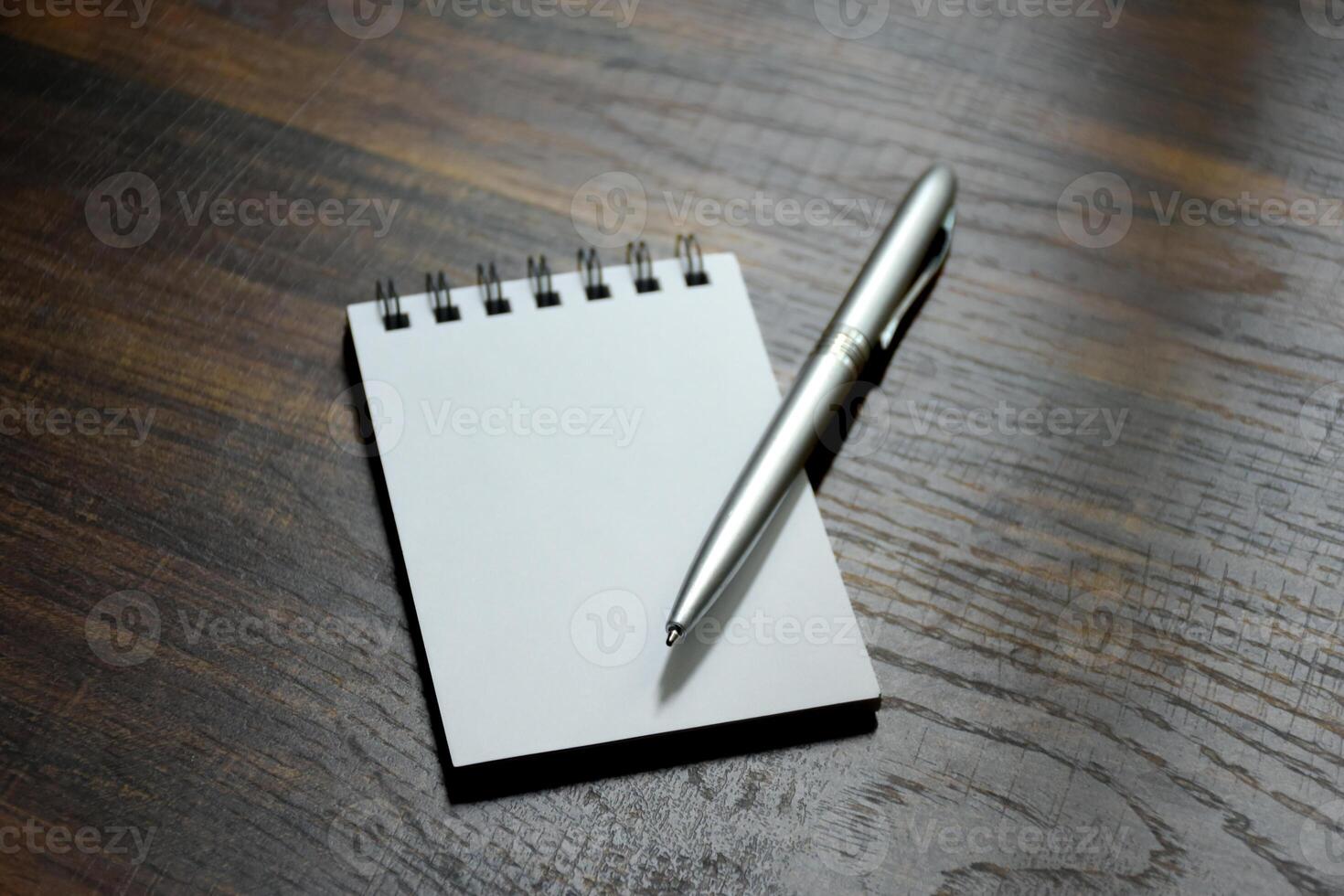 Blank paper notepad and silver pen placed on a beautiful wood-patterned desk at the office. photo