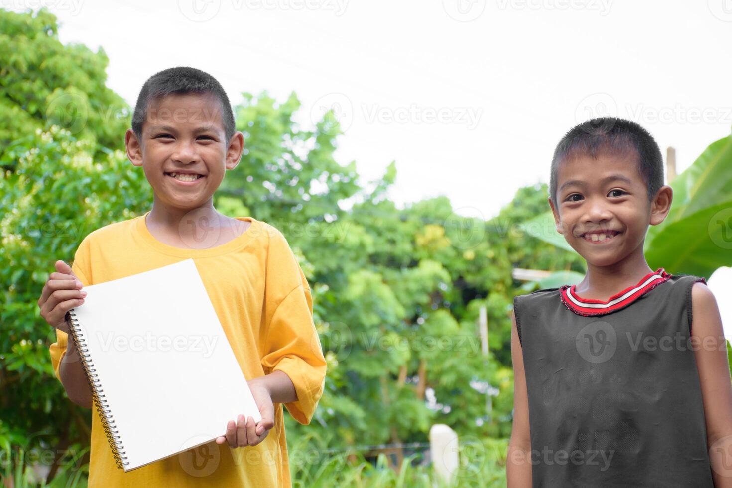 Young Asian two boys yellow and black t-shirt smiling and holding a blank white notebook on a blurred forest and tree background. photo