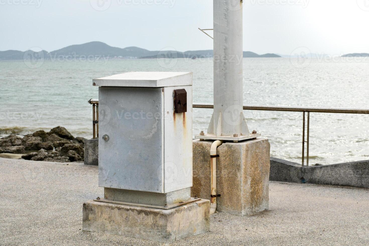 Old rusty electrical control cabinets or white control box and electrical poles sit on concrete floors on the seafront over mountain and sky cloudy background. photo