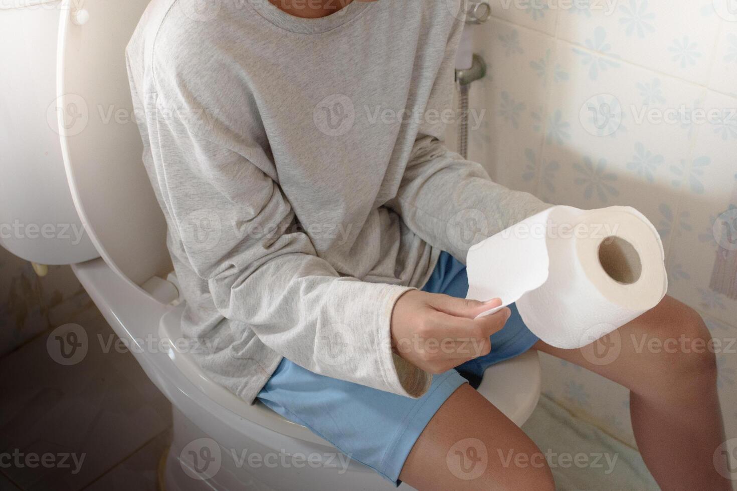Top-view Asian man sitting on the toilet bowl and his hand is using toilet paper or tissue in bathroom and soft sunlight blurred background for concept of excretion and hygiene. photo