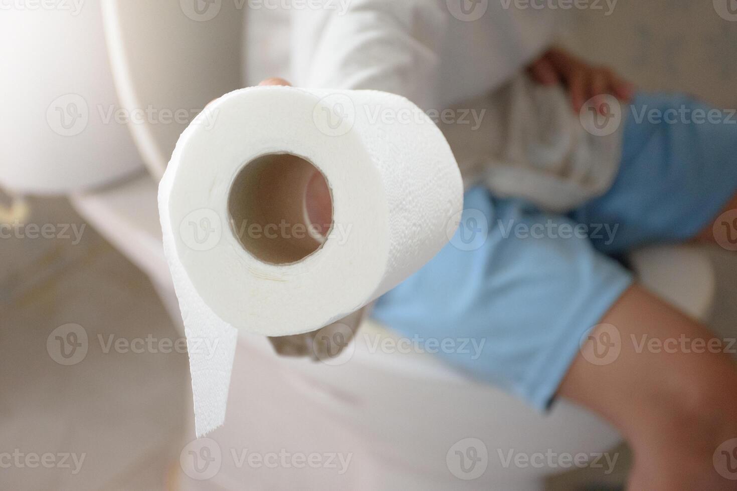 Man sitting on the toilet bowl and his hand is holding toilet paper or tissue close-up over blurred background in bathroom for concept of excretion and hygiene. photo
