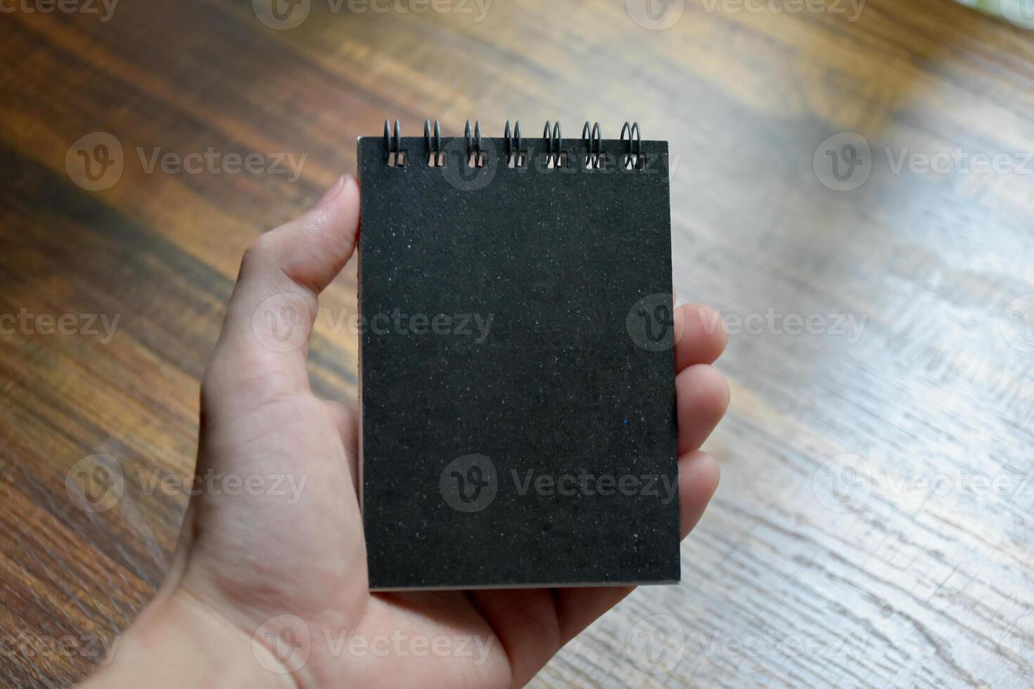 Hand holding a small black notebook on the background of a wooden patterned desk at the office. photo