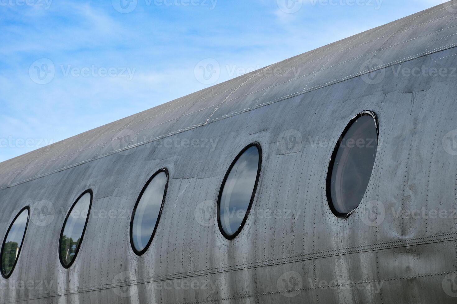 The body of the plane with circular windows shows a silver metallic surface of an idle airplane on the sky background. photo
