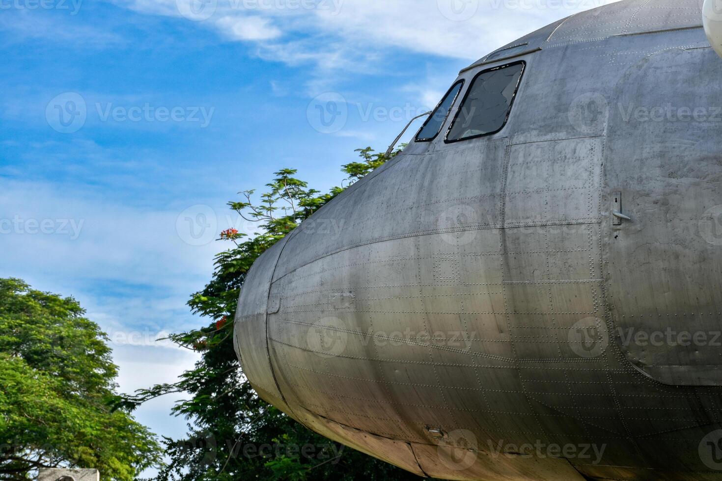 Front view of old airplane head part showing metal surfaces and broken window glass in tree forest and blue sky background. photo