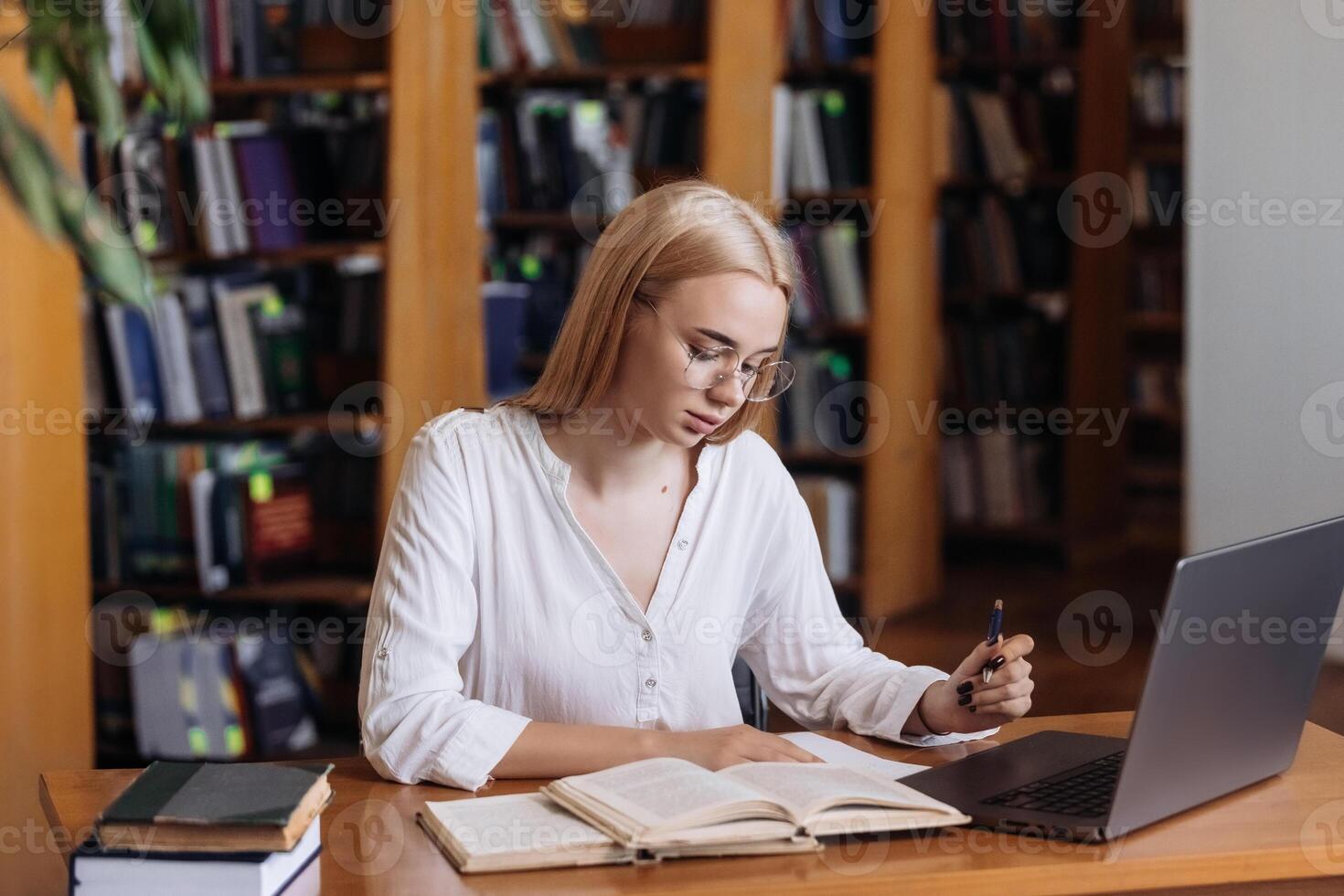 Serious hardworking blonde female student watching webinar, listening online learning course, looking at laptop display, writing notes at table in college library photo