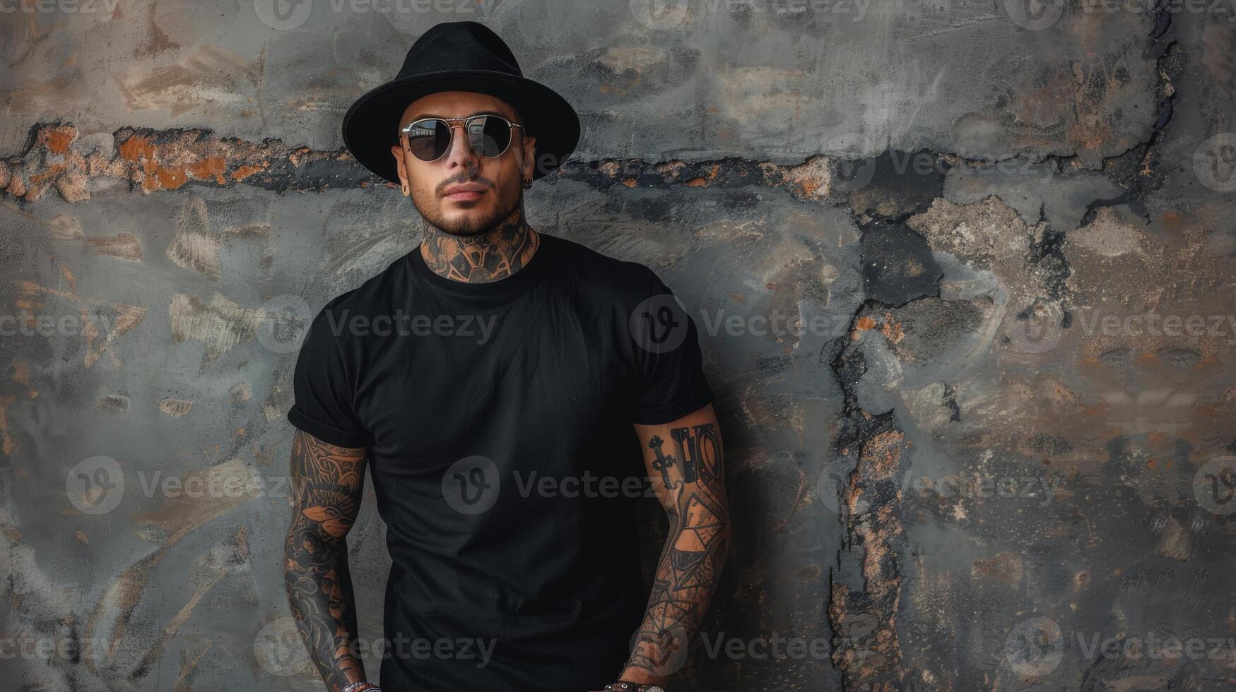 A young man with tattoos and a black fedora leans against a weathered wall photo