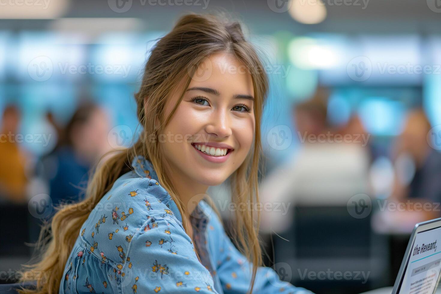 A young woman in a blue blouse is smiling while working on a laptop at an office desk photo