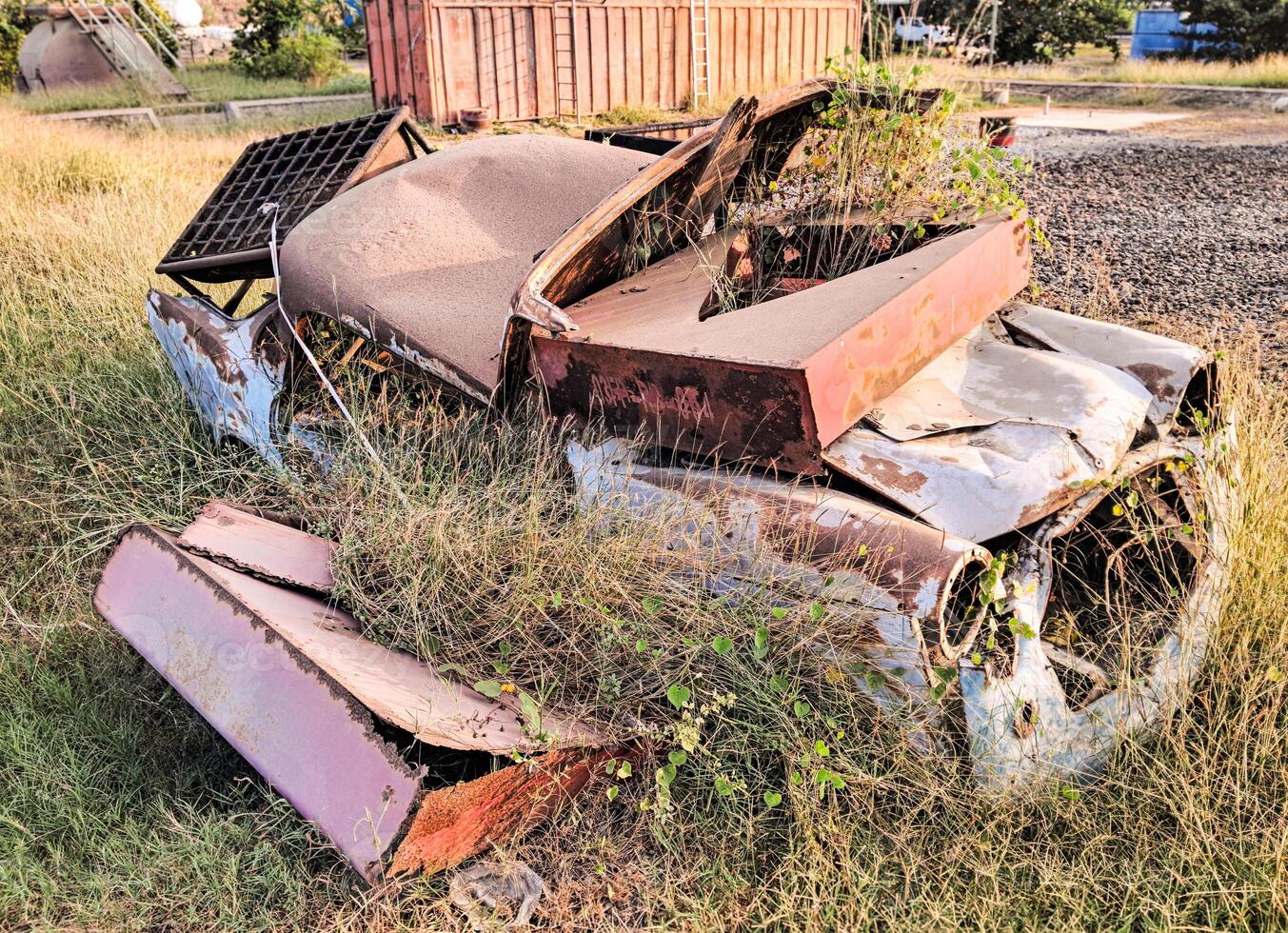 Broken abandoned car covered with rust in dry grassy field near tall trees against in Indonesian countryside photo