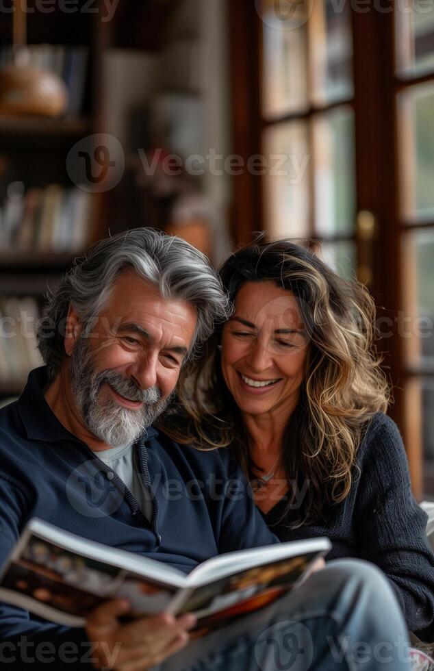 Joyful middle aged couple relaxes at home, laughing on sofa, sharing a magazine together photo