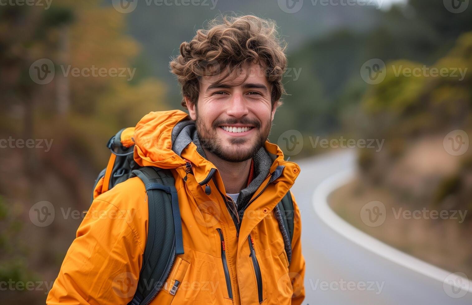 A cheerful man in an orange jacket stands on a road amidst lush greenery photo